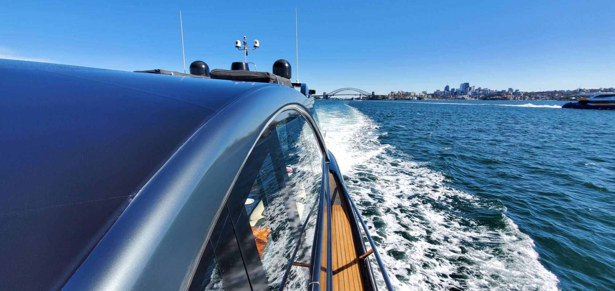 View from the side of a boat speeding through the water, leaving a trail of white waves. The city skyline is visible in the distance under a clear blue sky.