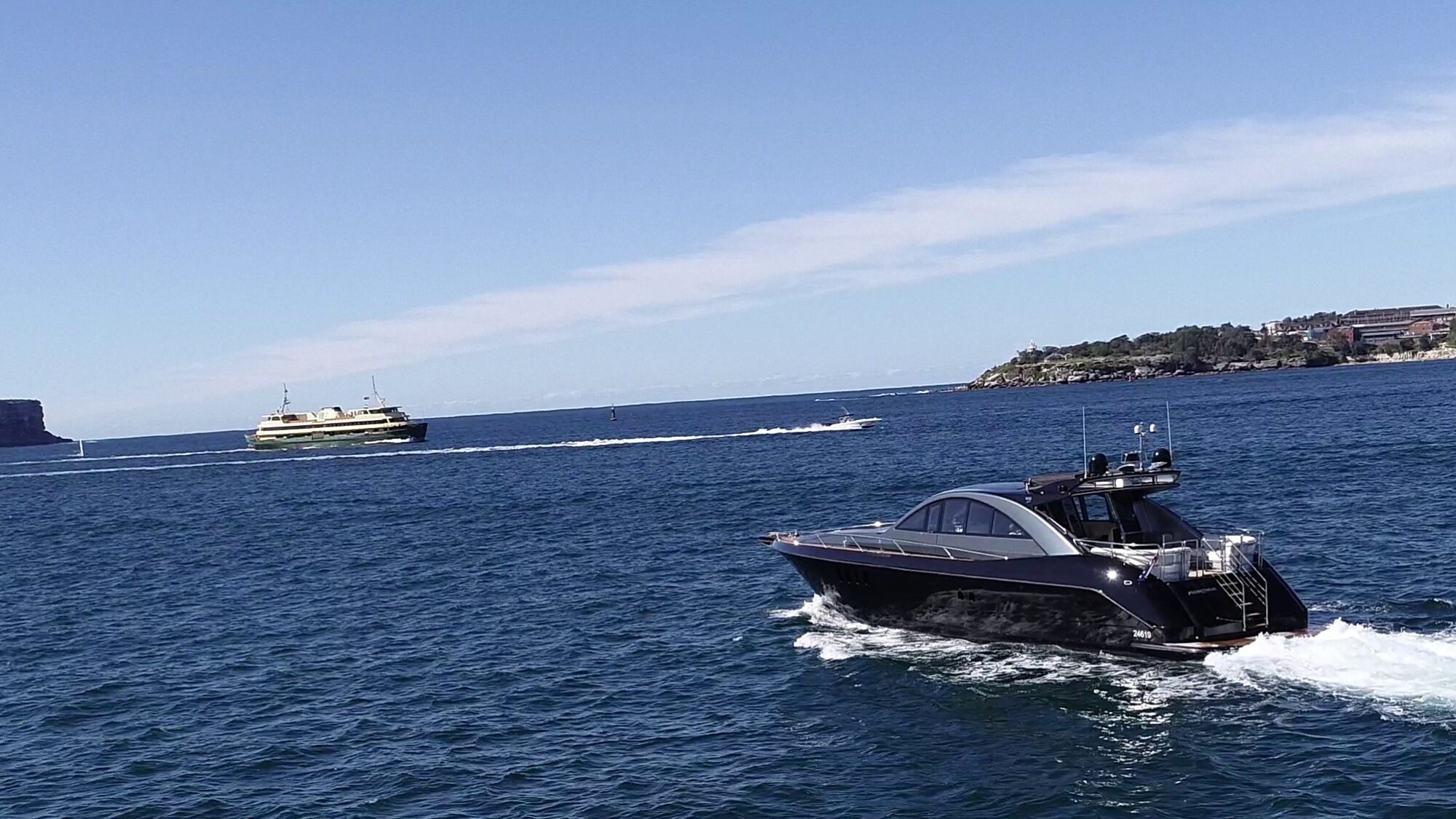 A sleek black motorboat cruises through the deep blue ocean, leaving a trail of white waves. In the background, a ferry moves across the water near a distant shoreline under a clear blue sky.