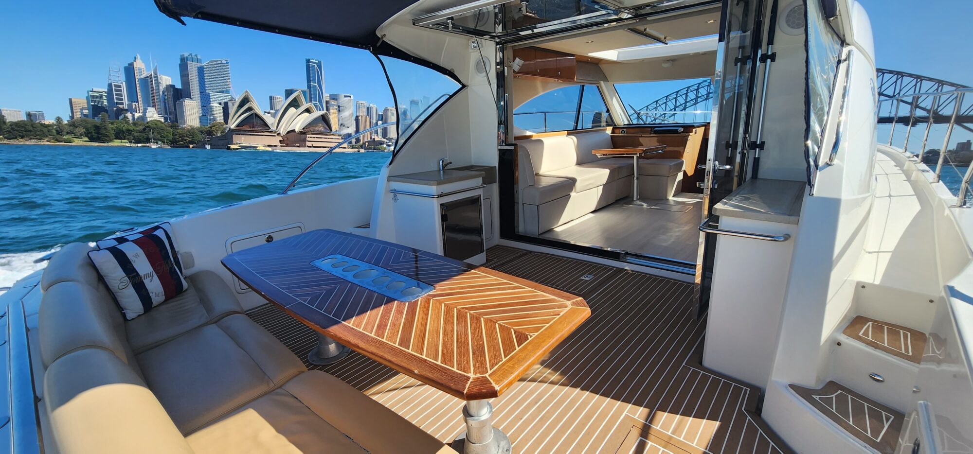 A luxurious yacht interior with polished wooden table and leather seating. The open deck offers a view of Sydney Harbour, featuring the Opera House, skyscrapers, and the Harbour Bridge under a clear blue sky.