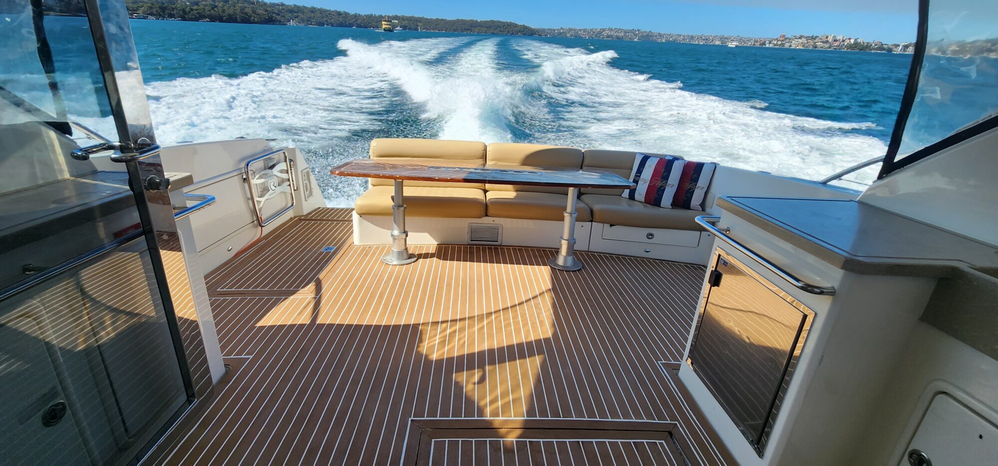 View from the back of a boat speeding over blue water, leaving a white wake behind. The deck has wooden flooring, a cushioned bench, a table with a blanket. The horizon shows green islands and a clear blue sky.