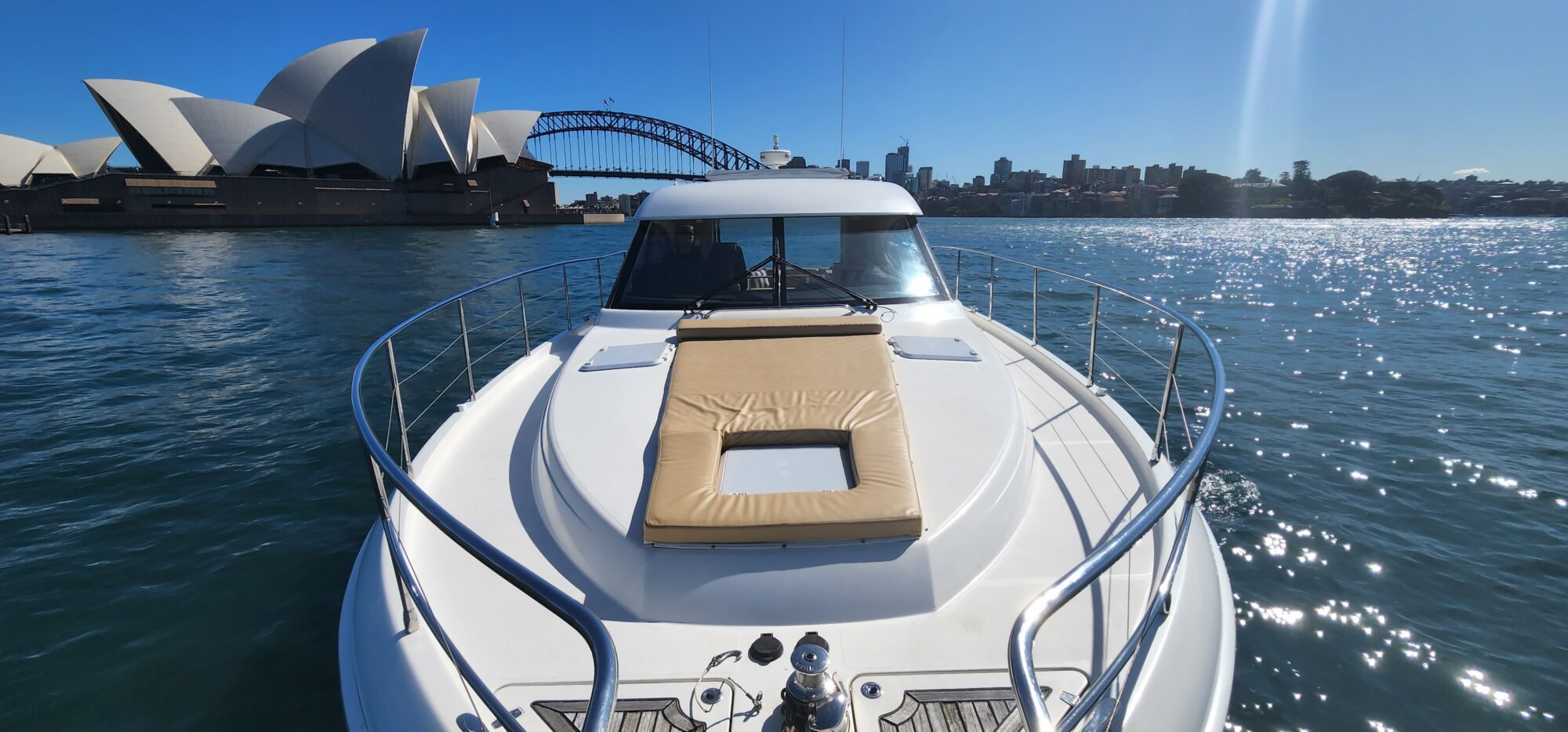 A white yacht cruises through Sydney Harbour, with the Sydney Opera House and Harbour Bridge visible in the background under a clear blue sky.