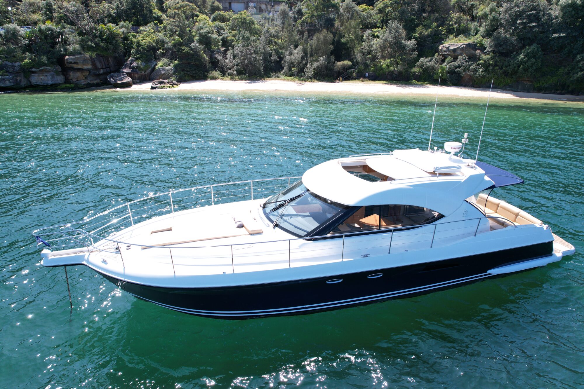 A sleek, white and black yacht floats on clear, green water. The boat has a streamlined design with a covered seating area. In the background, a small sandy beach is surrounded by lush green trees.