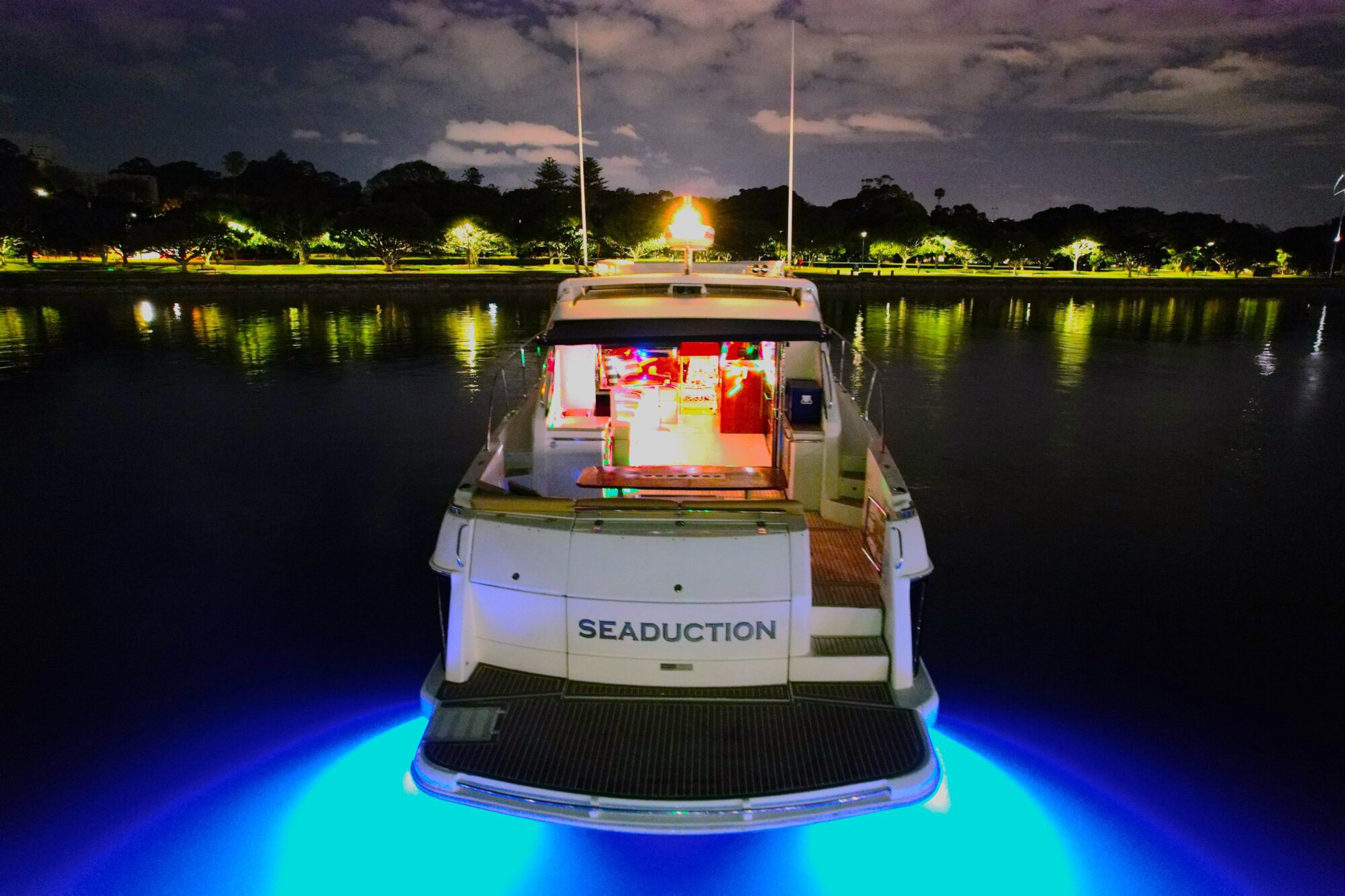 A yacht named "Seaduction" is illuminated by blue underwater lights at night. The boat is docked in calm waters, with a softly lit shoreline and silhouetted trees in the background under a cloudy sky.