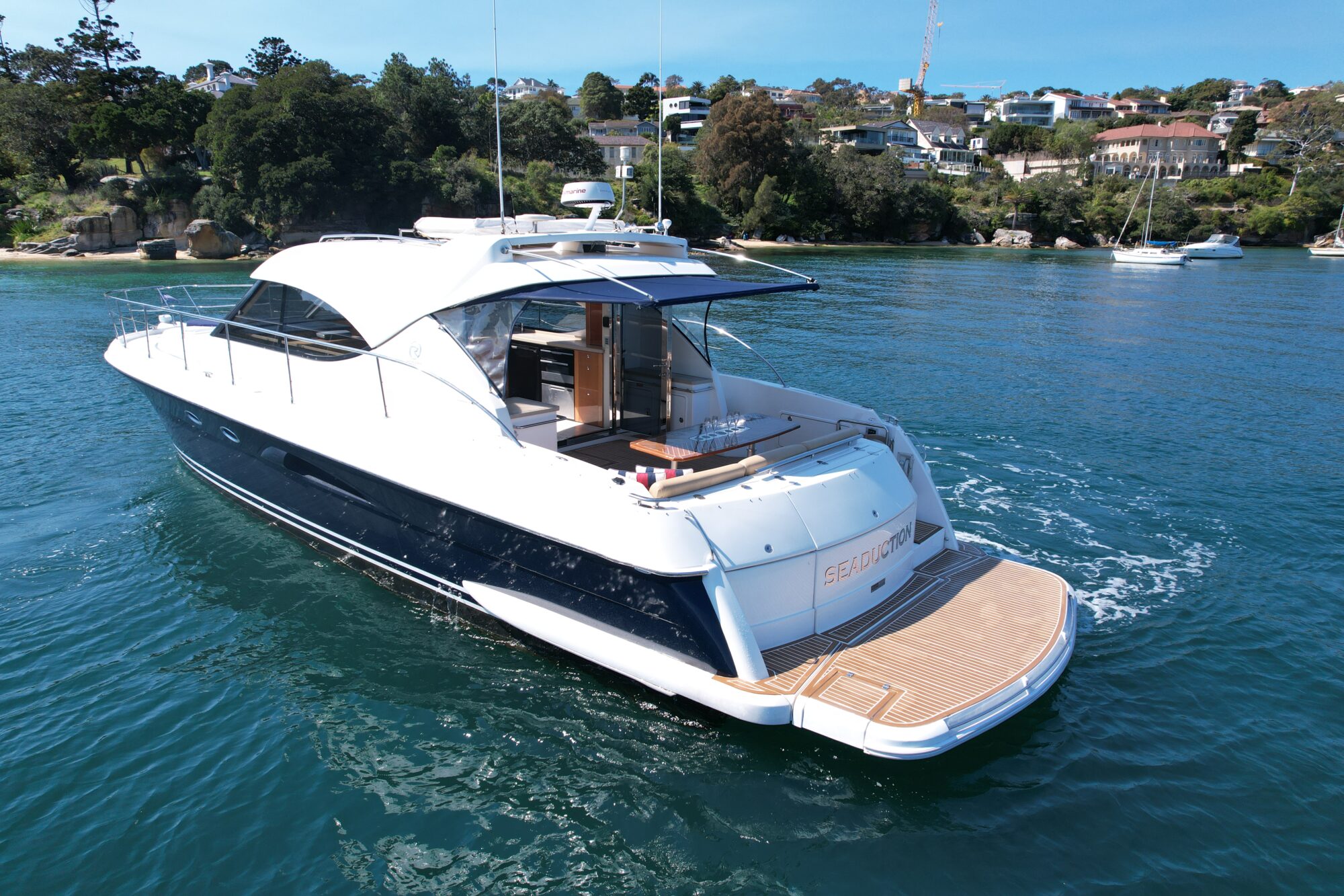 A sleek white yacht, named "SEDUCTION," sails on clear blue water. It features a spacious deck and open seating area. In the background are green trees and waterfront houses under a bright blue sky.