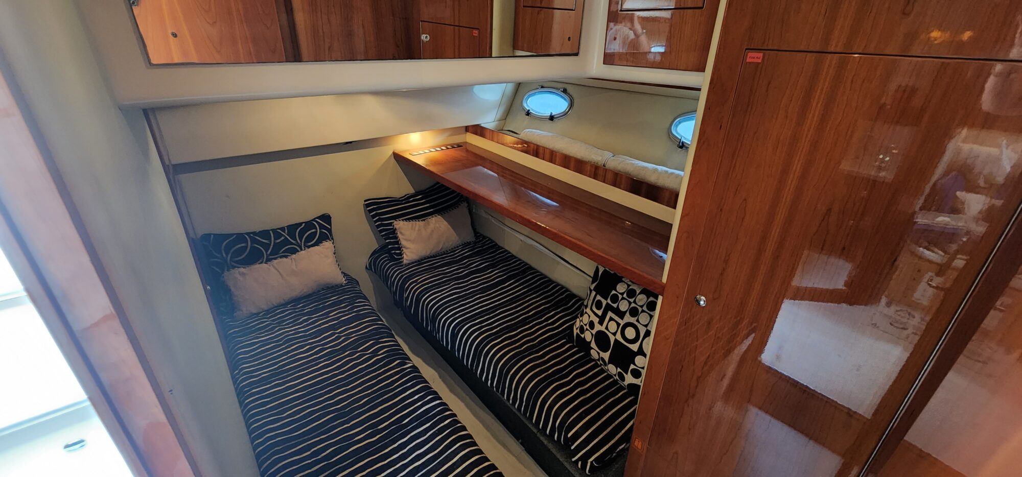 A cozy boat cabin with two twin beds featuring striped black and white bedding and pillows. The walls and shelves are wooden, with two small porthole windows and overhead lighting.