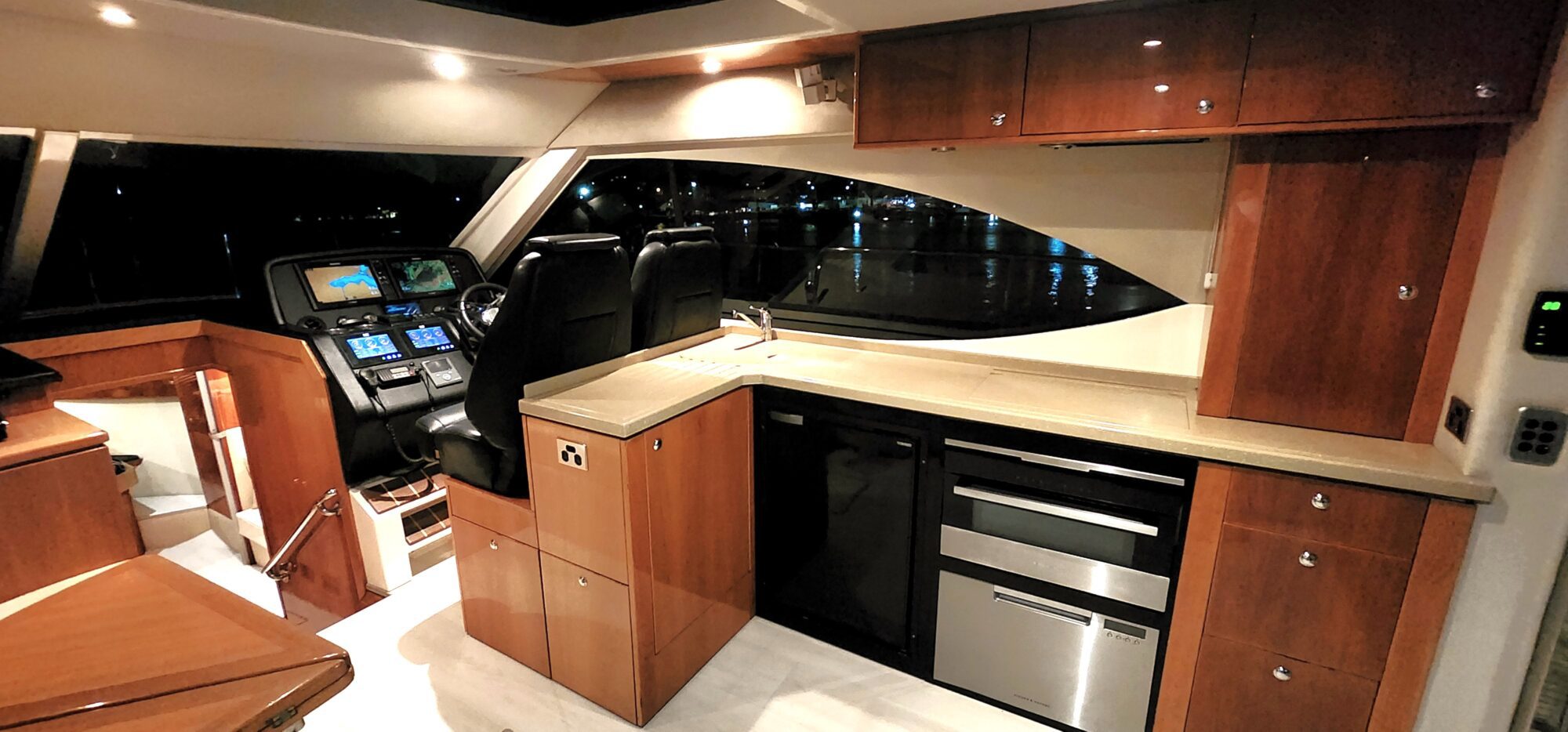 Luxurious yacht interior at night featuring a sleek kitchen with wooden cabinets, modern appliances, and a large window. The cockpit area with illuminated control panels is partially visible to the left.