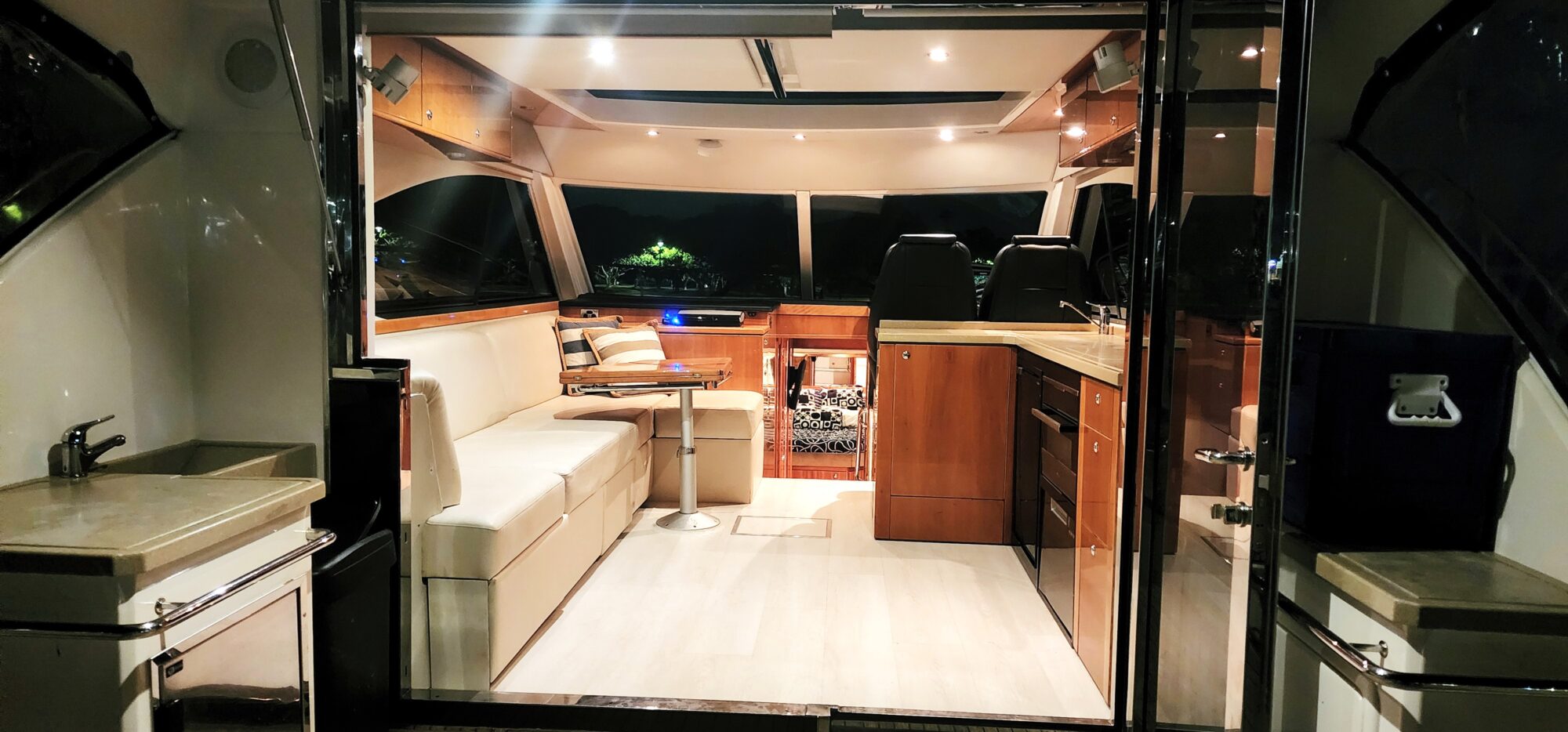 Interior of a modern yacht featuring a cozy seating area with a beige couch, a small table, and two black captain's chairs. The warm lighting highlights wood accents and a kitchenette on the right with a sink and mini fridge.