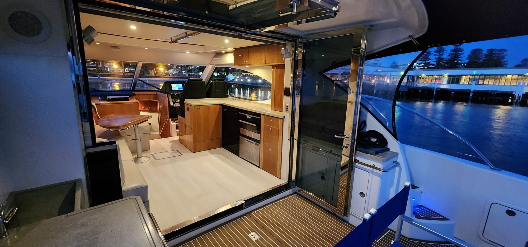 Interior of a boat at night, featuring a modern kitchen with wooden cabinets, a small dining area, and glass doors. The view outside shows a calm waterfront with reflections of city lights.