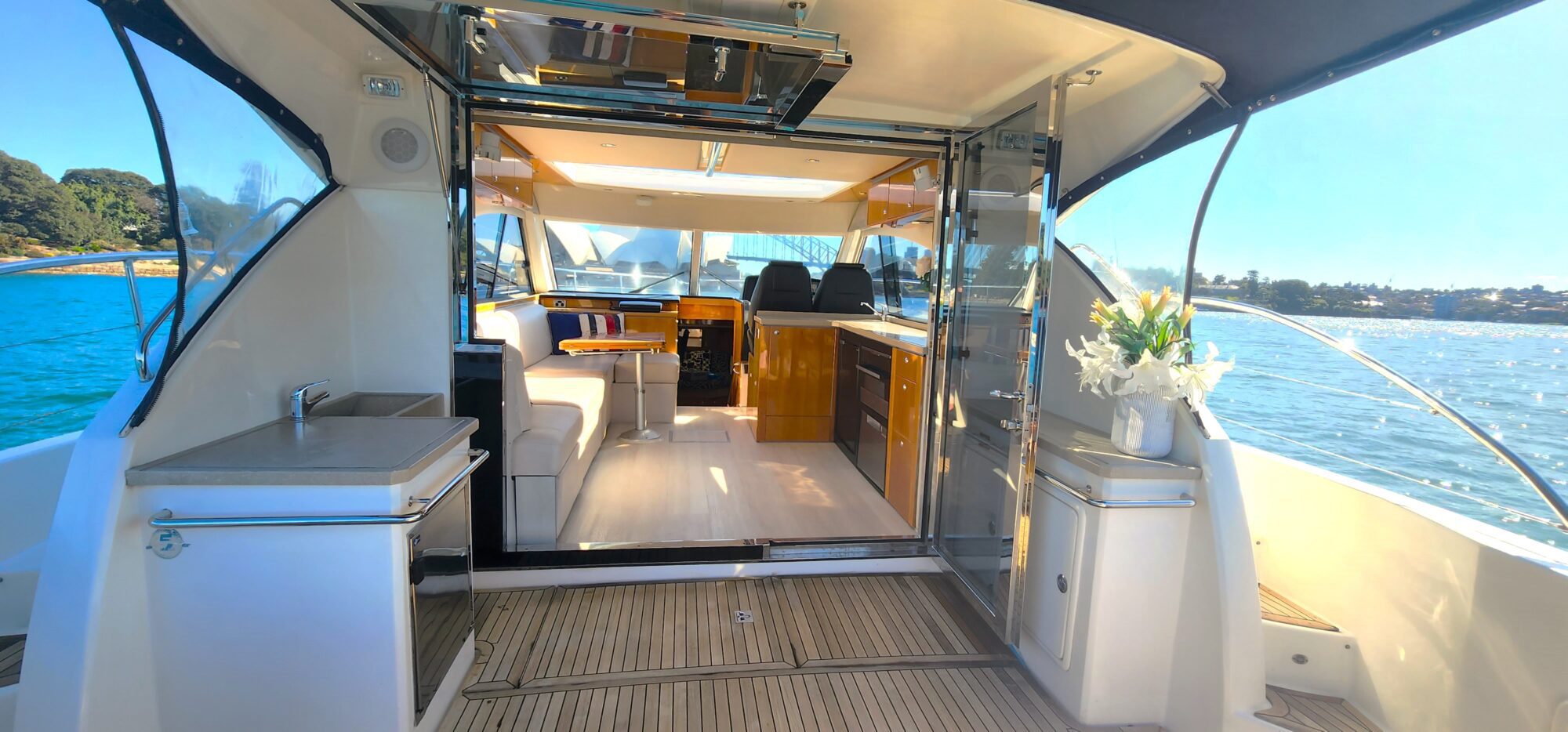 View of a luxurious yacht interior from the deck, featuring elegant wooden finishes, white cushions, and a vase of flowers. The background shows calm blue water and distant greenery under a clear blue sky.