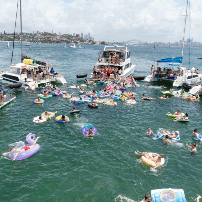 A lively scene on a sunny day with groups of people on boats and inflatables enjoying a party on the water. In the background, a cityscape with tall buildings and a bridge is visible. The water is dotted with colorful floaties and swimmers.