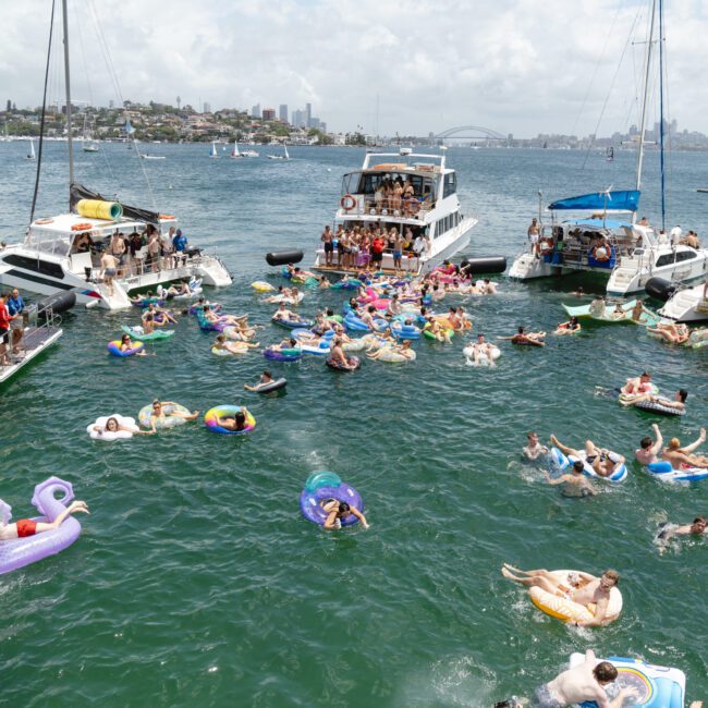 A lively scene of people swimming and relaxing on colorful inflatables in the water near several boats. The sky is partly cloudy, and a city skyline is visible in the background.