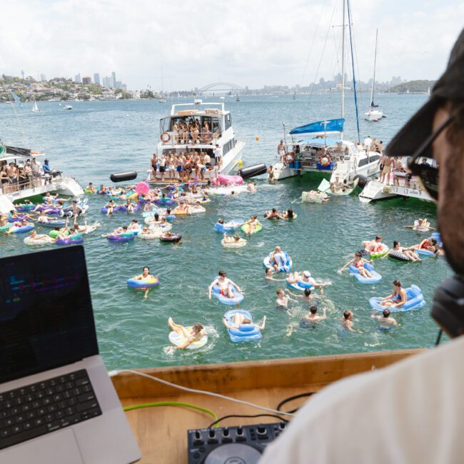 A DJ performs on a boat overlooking a group of people floating on inflatables in the water. Several boats are anchored nearby, and a city skyline is visible in the distance under a partly cloudy sky.