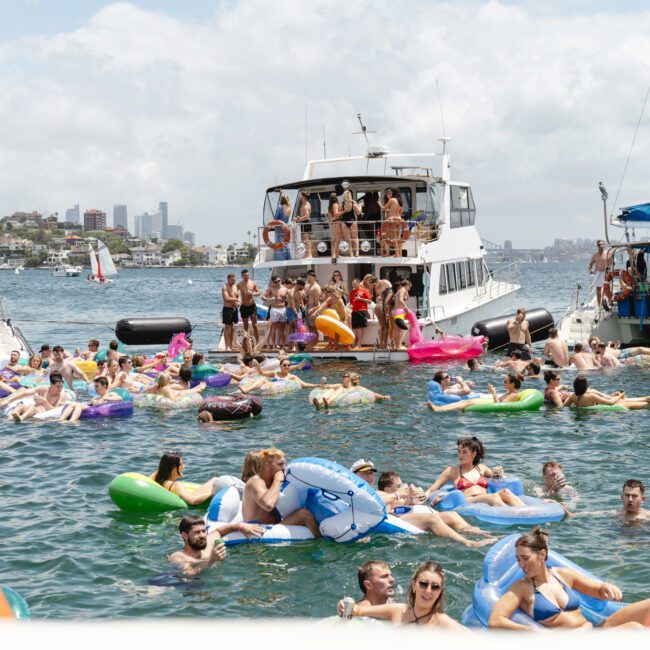 A lively scene with a crowd of people on colorful inflatable rafts and floats enjoying a sunny day on the water. Boats are docked nearby, and the cityscape is visible in the background under a partly cloudy sky.