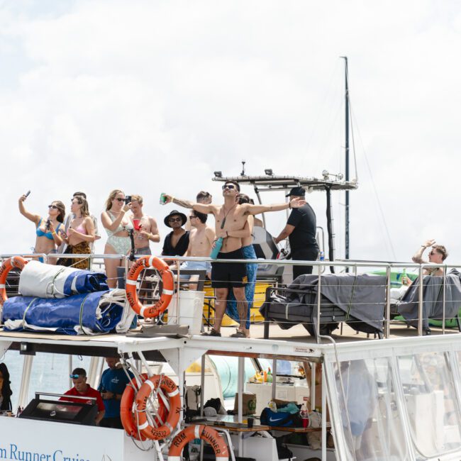 A group of people on a boat enjoying a sunny day. Some are sunbathing on the upper deck, while others are taking photos and socializing. The boat is on clear water, and life preservers are visible along the sides of the vessel.