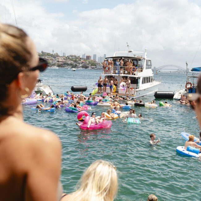 A crowded boat party with people swimming in the ocean on inflatable pool toys. The water is filled with colorful floaties, and a large boat in the background hosts more partygoers. The sky is partly cloudy.