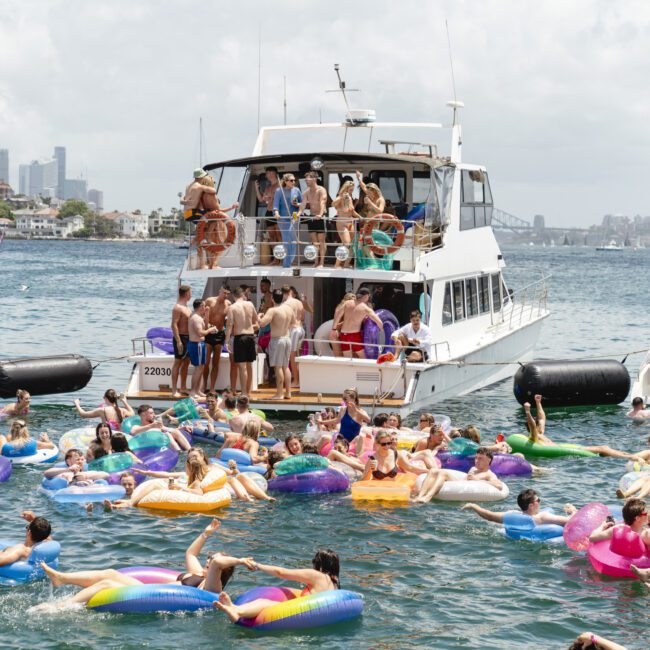 A large group of people are enjoying a sunny day on a boat and in the water. Many are on colorful inflatable floats near the boat, with a city skyline in the background. The scene is lively and festive.