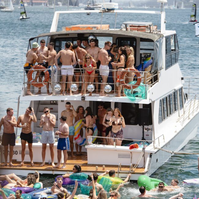 A crowded boat party with people in swimsuits enjoying the sunny weather on two levels. Others float on colorful inflatables in the water. In the background, a bridge and cityscape are visible.