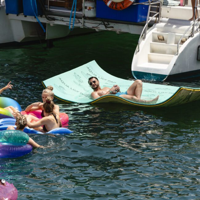 People relaxing in a body of water, floating on colorful inflatables and a large green mat. A few boats are docked nearby. The scene is lively and sunny, suggesting a fun, social atmosphere.