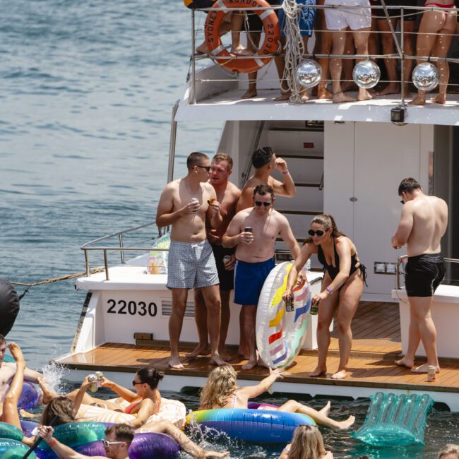 A group of people enjoying a sunny day on a yacht, with several holding drinks. Many others are in the water on colorful floats near the boat. The scene is lively and festive against a backdrop of blue water.