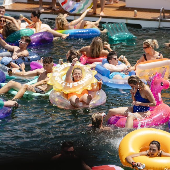 A group of people enjoying a sunny day in the water, surrounded by colorful inflatable pool floats shaped like unicorns, donuts, and other fun designs. A boat is visible in the background.