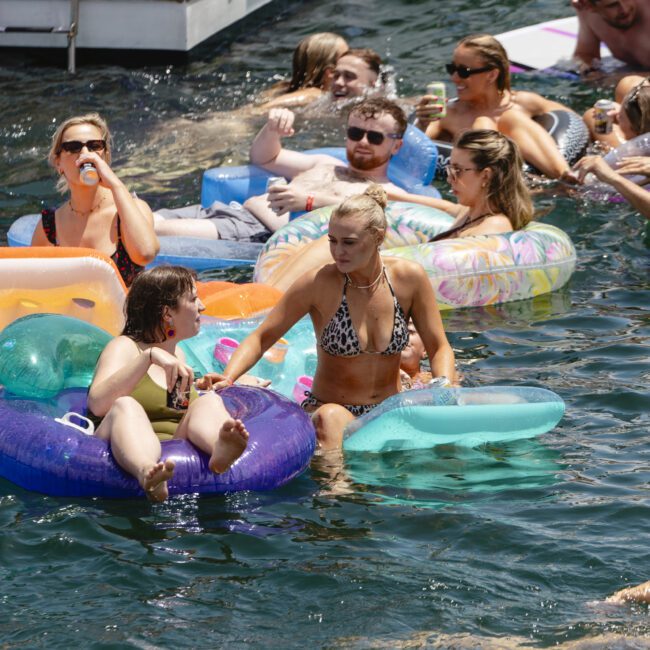 A group of people relax on colorful inflatable tubes in the water near a boat. They are enjoying drinks and the sunny day. In the background, more people can be seen lounging on the boat and in the water.