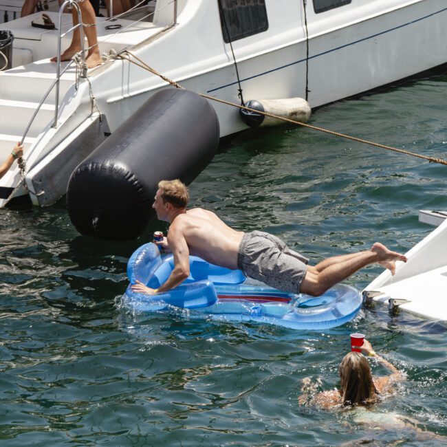 A person in a swimsuit kneels on an inflatable pool lounger, balancing it between a boat and the water. Another person swims nearby holding a cup, while a third person on a purple floater is also visible in the water.