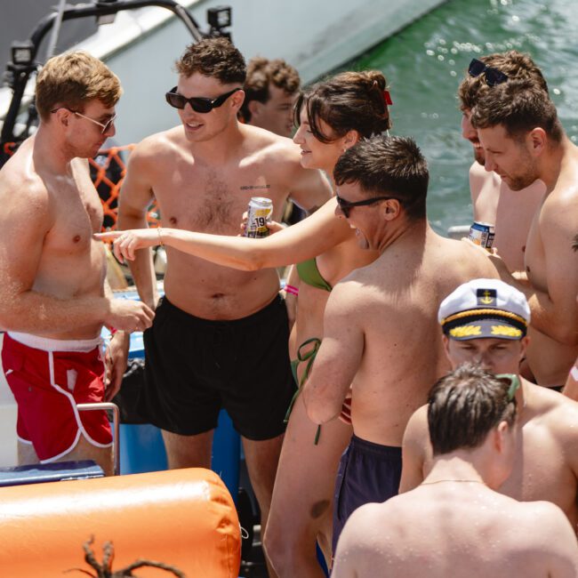 A group of people in swimsuits having fun on a boat. Some are holding drinks, and one person in the front is wearing a captain's hat. The water is visible in the background, indicating they are on a lake or ocean.