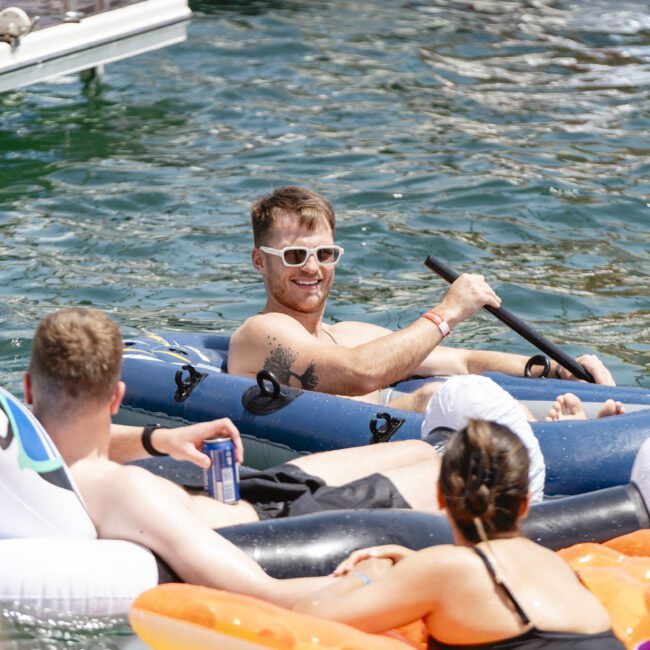 A group of people are relaxing on inflatable floaties in a sunny pool or lake. One person wearing white sunglasses is paddling on a blue float. Others are lounging with drinks, and the scene is lively and cheerful.