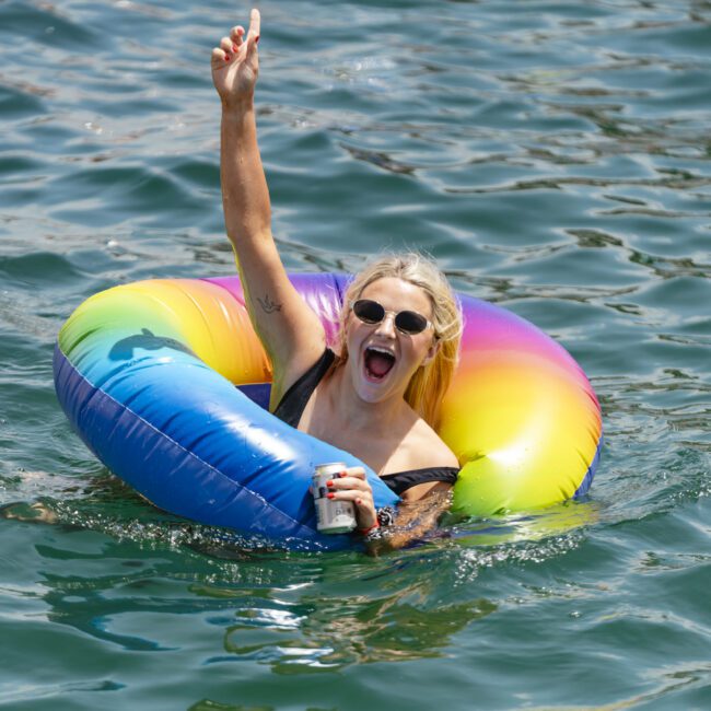 A person wearing sunglasses and a black swimsuit is smiling and raising one arm while sitting in a rainbow-colored inflatable ring on the water. They are holding a can in the other hand.