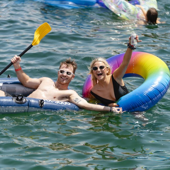 Two people are floating in a lake, smiling and holding up beverages. One person is in an inflatable boat with a paddle, and the other is in a rainbow-colored inner tube. The water is green and they appear to be enjoying the sunny day.