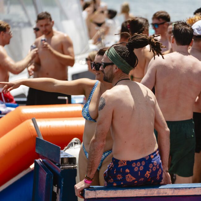 A group of people in swimwear socializing on a boat. Some are standing and talking, while others are sitting near inflatable floaties. The atmosphere is relaxed and sunny. In the background, the sea and part of another boat are visible.