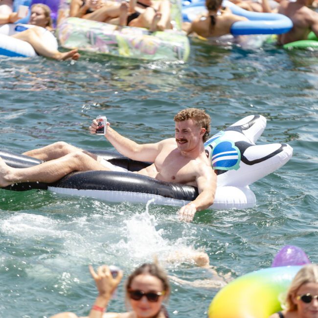 A shirtless man with a mustache relaxes on an inflatable black and white float shaped like a dog, holding a drink. He's surrounded by others on colorful inflatables enjoying the water on a sunny day.