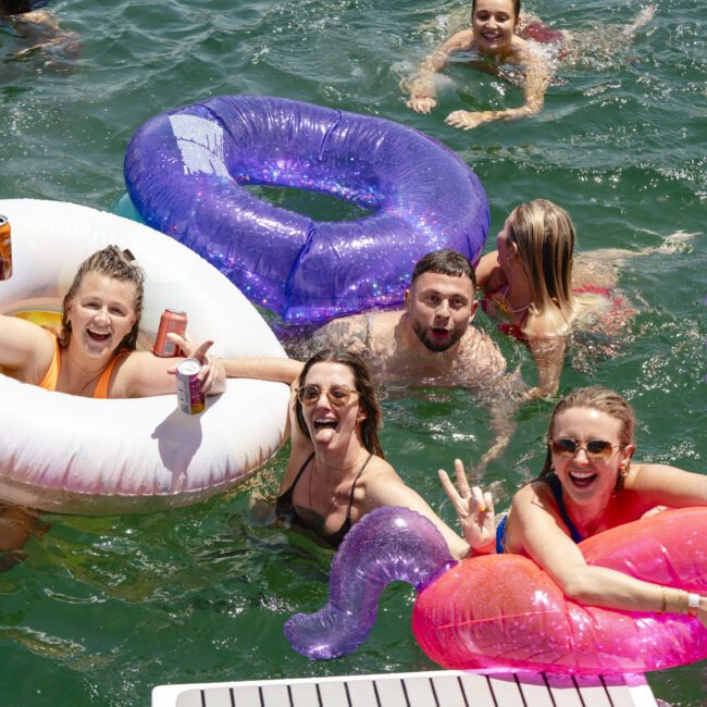 A group of people enjoying a sunny day in a lake, floating on colorful inflatable rings. Some are holding drinks and smiling at the camera, with others swimming in the background. The water is a vivid green.