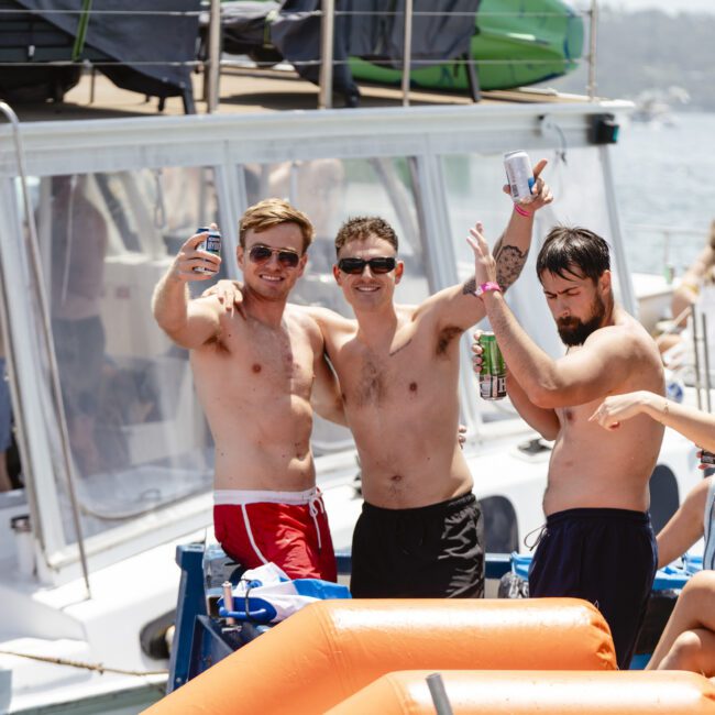 A group of people enjoying a sunny day on a boat. Three men in swim trunks hold drinks and pose playfully for the camera, while others socialize nearby. The boat is on a lake or the sea, with trees visible in the background.