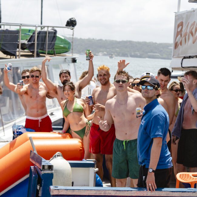 A group of people in swimwear enjoy drinks on a boat under sunny skies. Some are sitting or standing near the boat's railing, while others wave at the camera. A crew member in a blue shirt is in the foreground. The ocean and another boat are visible.