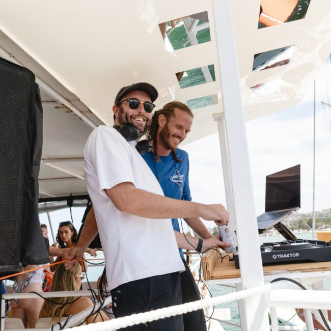 Two DJs set up equipment on a boat deck. One wears a white shirt and sunglasses, smiling, while the other in a blue shirt adjusts controls on a laptop. People in the background enjoy the sunny day on the boat.