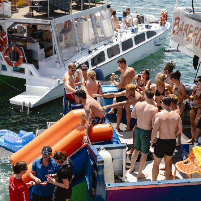 A lively scene of people enjoying a sunny day on the water. Two boats are docked close together, with groups of people socializing, relaxing on inflatable floats, and preparing to swim. The water is calm and a few boats are visible in the background.