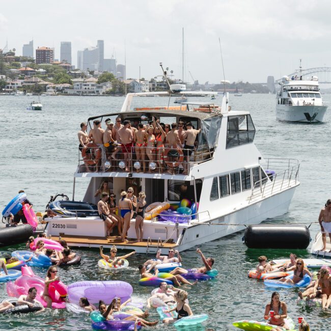 A group of people enjoying a party on a yacht in a harbor, surrounded by others floating on colorful inflatables in the water. The city skyline and a bridge are visible in the background under a partly cloudy sky.