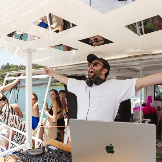 A DJ in a white shirt and cap stands on a boat with arms outstretched, appearing joyful. People around are enjoying themselves, some dancing. The scene is set on a sunny day with water and greenery in the background.