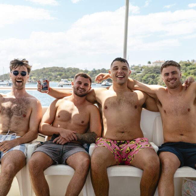 Four men sit on a boat, smiling and holding drinks, with scenic views of water and greenery in the background. They appear relaxed and cheerful under a sunny sky.