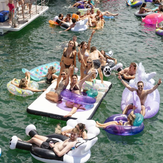 A group of people enjoy a sunny day on the water, lounging on colorful inflatable pool floats. They are smiling, holding drinks, and some are waving. The scene is lively and festive.