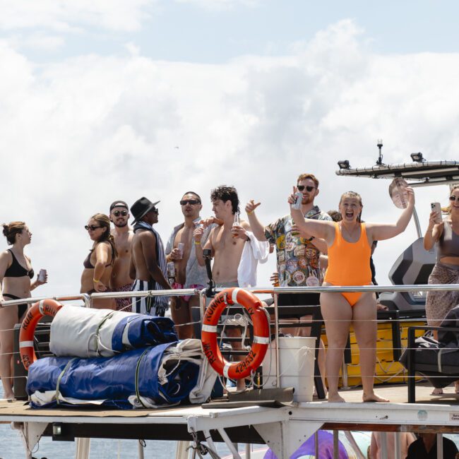 A group of people enjoying a sunny day on a boat. They are laughing and holding drinks, wearing swimwear and summer outfits. The boat is equipped with life preservers and equipment. The background shows a cloudy sky and distant shoreline.