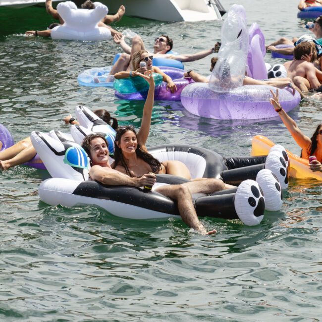 A group of people enjoying a sunny day on the water, relaxing on colorful pool floats of various shapes, including animals. They are smiling, raising their arms, and appearing to have a good time.