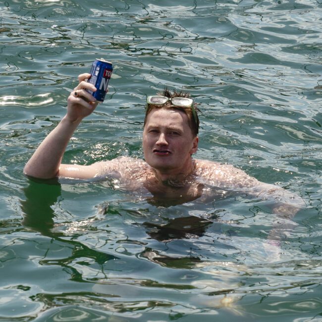 A man in water holds up a blue can, wearing sunglasses on his head. The water around him is rippling, and he appears to be enjoying a swim on a sunny day.