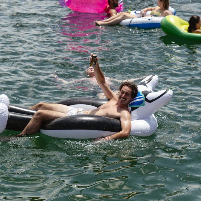 A man lounges on an inflatable float in the water, holding up a drink and smiling. He is surrounded by other people on colorful floats enjoying a sunny day on the lake.