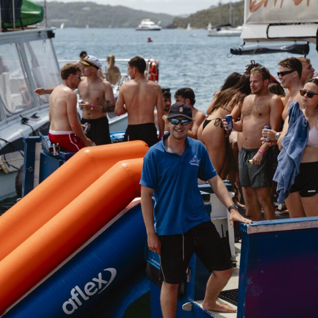 A group of people in swimwear enjoy a boat party. A man in a blue shirt and sunglasses stands smiling on the boat's steps, with inflatable slides and a "bar" sign in the background. The scenic backdrop includes water and hills.