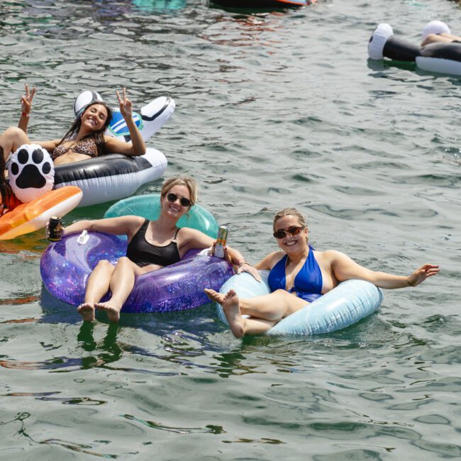 People are relaxing on colorful inflatable floats in a body of water, enjoying a sunny day. Two women in the foreground are smiling, wearing sunglasses and swimsuits, and appear to be having fun.