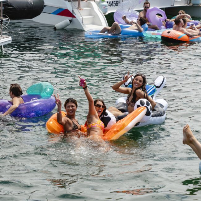 A group of people enjoy a sunny day on the water, floating on colorful inflatable rafts near a boat. Some are relaxing while others take photos, all surrounded by clear, rippling water.