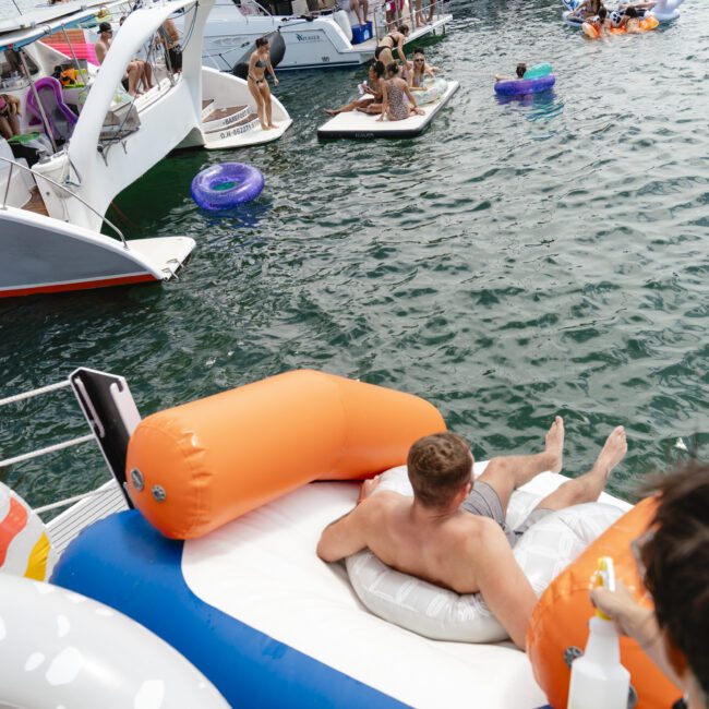 A group of people enjoying a day on the water. Several boats are docked closely, with people swimming and lounging on inflatable rafts. The scene conveys a fun and relaxing atmosphere.