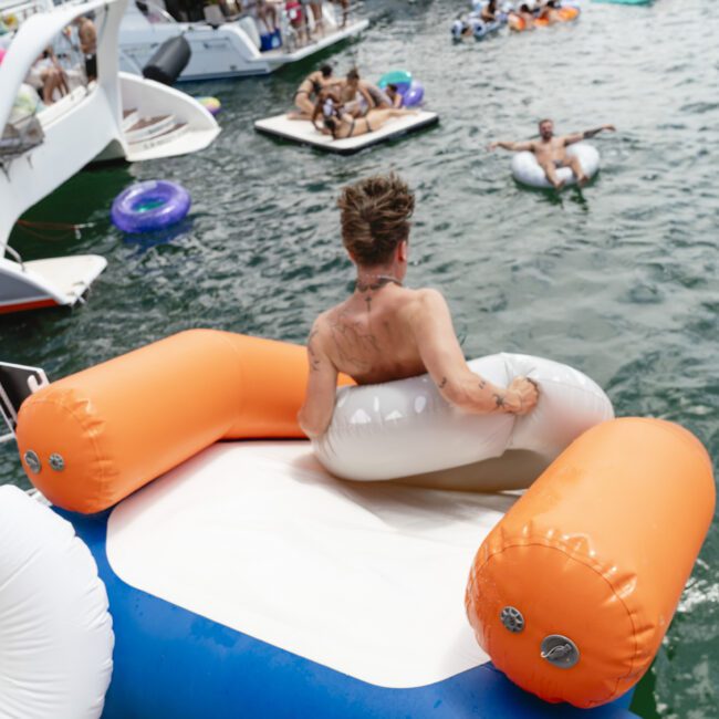 A person is about to slide from an orange and blue inflatable slide into a body of water. Other people are lounging on inflatable tubes nearby, with boats in the background. The scene conveys a lively and fun atmosphere.